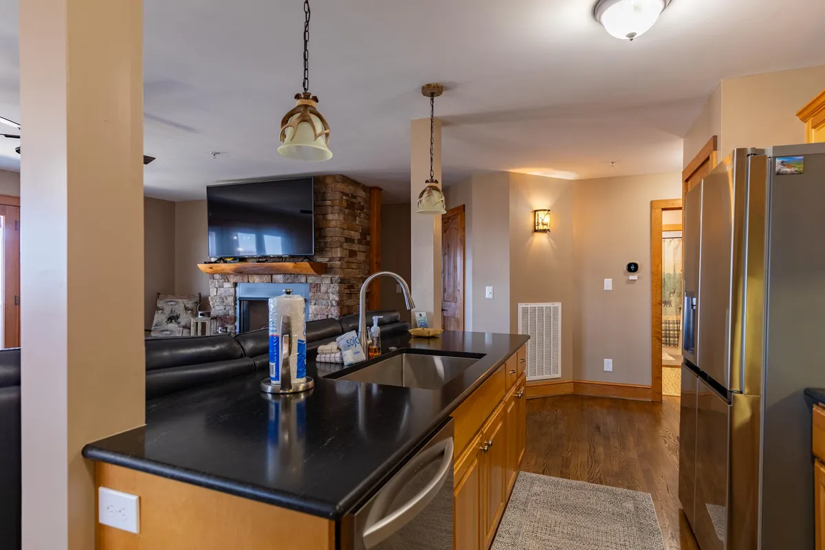 A cozy perspective highlighting the kitchen island with a black countertop and wooden cabinets, adjacent to the stone fireplace and television. The layout offers a glimpse of the kitchen’s functionality and its connection to the living space.