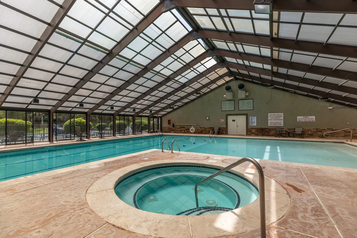 An inviting indoor swimming pool area featuring a large glass roof that fills the space with natural light. The pool is complemented by a bubbling hot tub, with plenty of seating for relaxation and socializing. The modern design and tranquil atmosphere make it ideal for both exercise and unwinding.