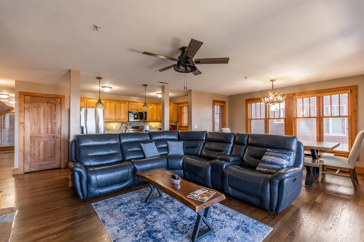 A wide view of the open-concept main living area, showing a large leather sectional sofa in the foreground, a wood coffee table, and a chandelier above the adjacent dining area. The kitchen with stainless steel appliances and wood cabinetry is visible in the background, seamlessly connecting the living, dining, and kitchen spaces.