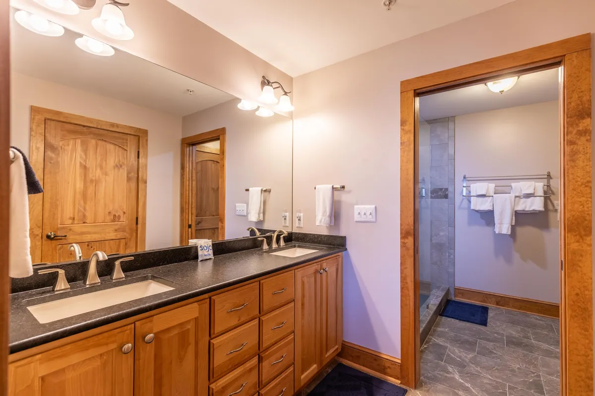 A spa-inspired bathroom featuring a double sink vanity with sleek wooden cabinetry, a large mirrored wall, and a custom-tiled shower area. Morning light streams in, enhancing the calm, high-end feel of the space.