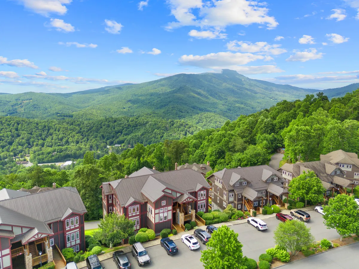 The image showcases a row of charming houses nestled amid lush greenery, with sweeping mountain vistas under a dramatic, cloudy blue sky. The scene exudes tranquility, with the inviting porch promising stunning sunset views and opportunities for relaxation and wildlife watching.