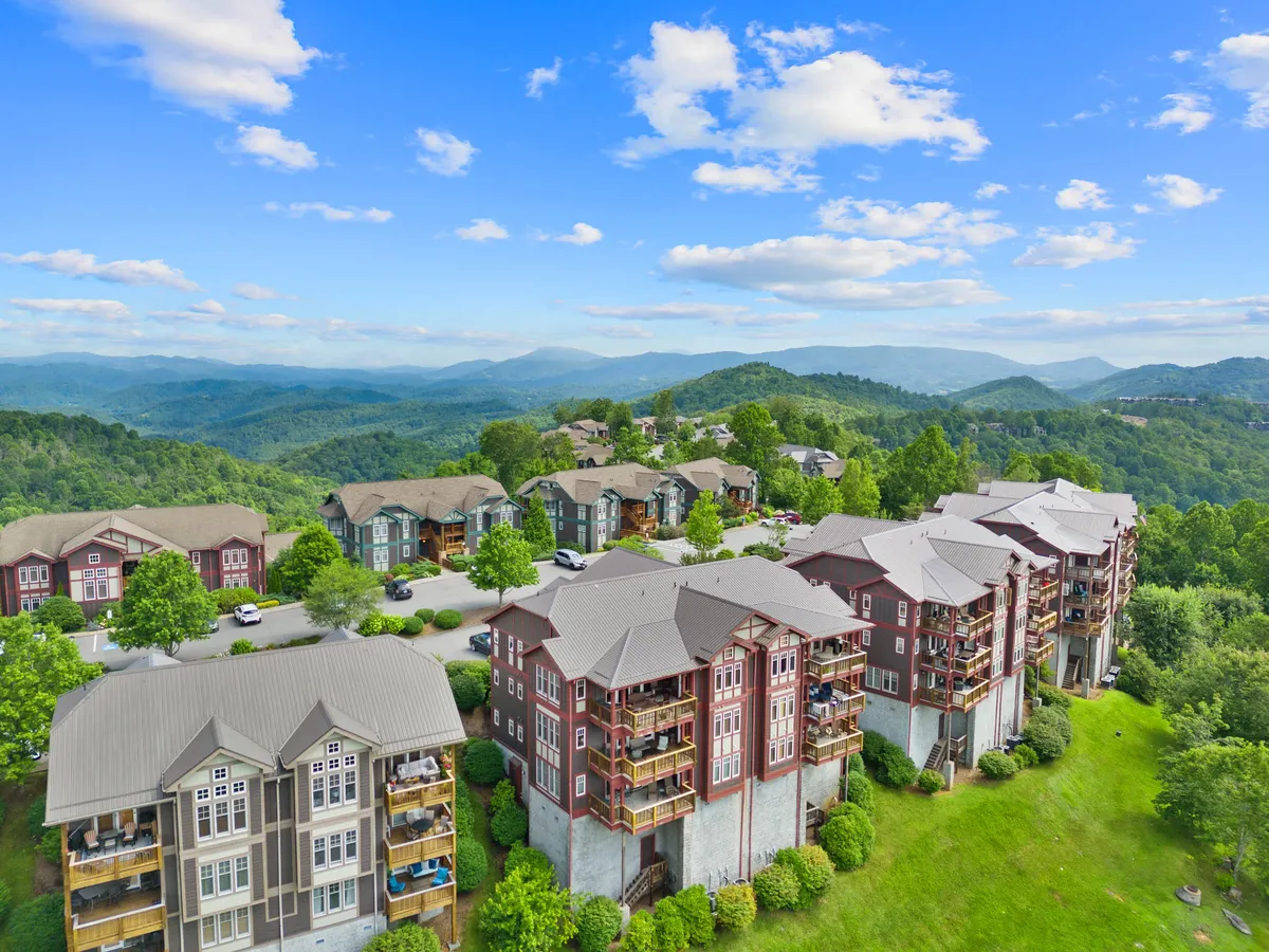 A scenic exterior shot of the apartment buildings nestled against lush green hills with a dramatic mountain backdrop and a blue sky dotted with clouds. Balconies face out towards the landscape, inviting residents to enjoy the breathtaking views and fresh air.