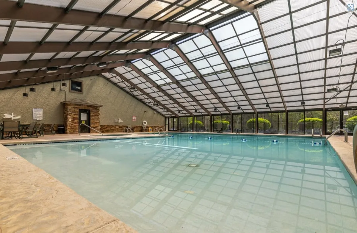 An inviting indoor swimming pool sits beneath a large glass ceiling, with comfortable seating arranged along the sides. The light-filled space suggests opportunities for rejuvenation, relaxation, and socializing at the nearby resort clubhouse.