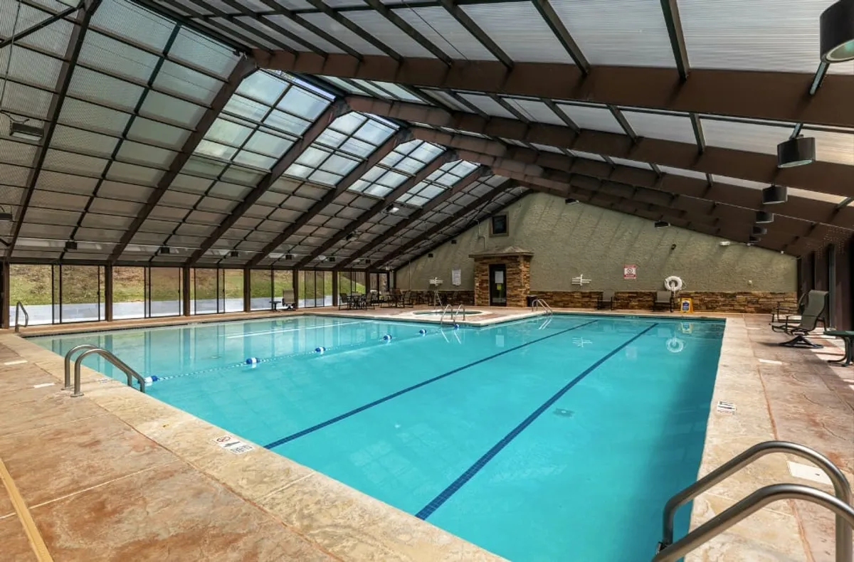 An interior view of a spacious indoor swimming pool area, bathed in natural light from a large glass roof and surrounded by seating. The tranquil atmosphere offers a glimpse into the relaxation and recovery options available after outdoor activities.