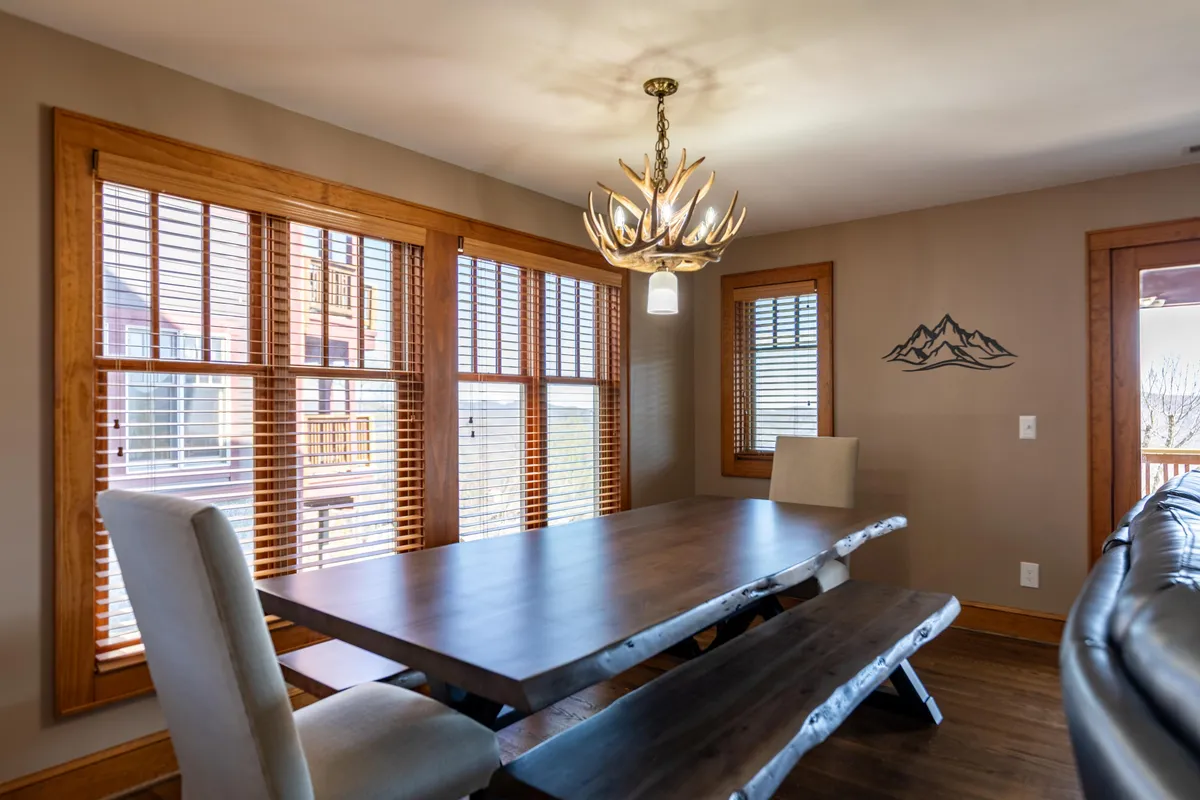 A close-up of the dining area featuring a large wooden live-edge table beneath a statement antler chandelier. Wooden blinds and mountain-themed artwork add to the warm, inviting ambiance, with ample space for family meals and gatherings.