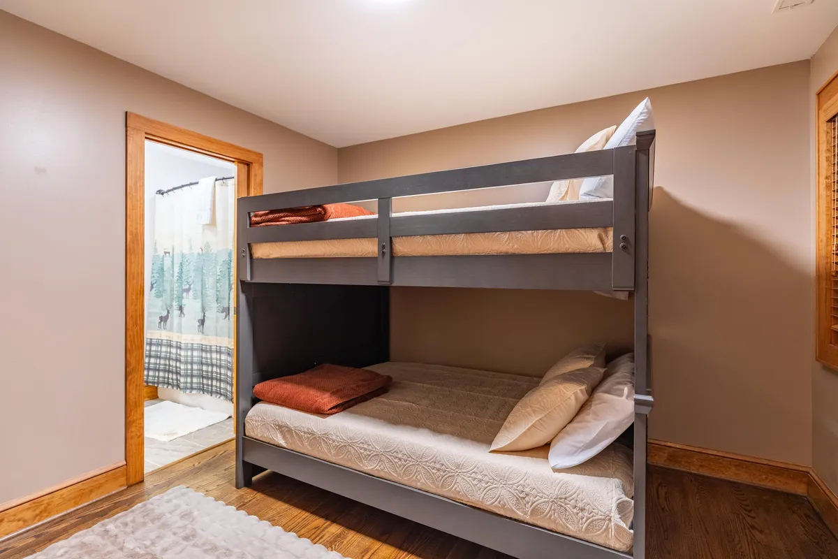 A cozy bunk bedroom with a full-over-full bunk bed dressed in decorative pillows, adjacent to a doorway that leads to a private bathroom. The room’s layout is welcoming for families or children, emphasizing both functionality and comfort.